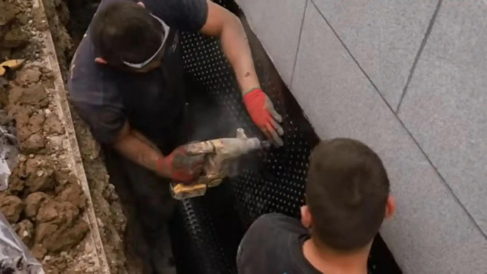 Workers installing exterior foundation drainage membrane in an excavated trench