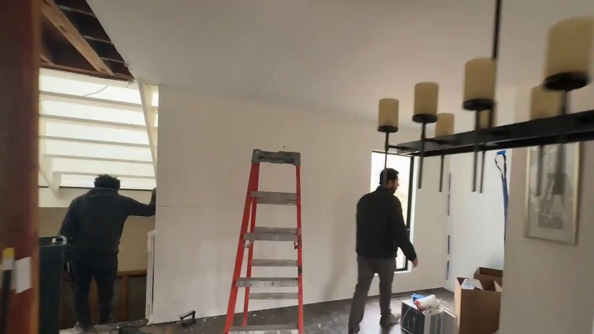 Interior renovation in progress with exposed ceiling framing, a ladder, and workers repairing a water-damaged room